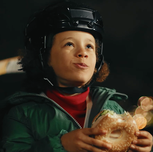 Child in hockey gear eating a Dempster's® bagel sandwich