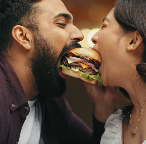 A happy couple, humorously taking a bite from the same sandwich