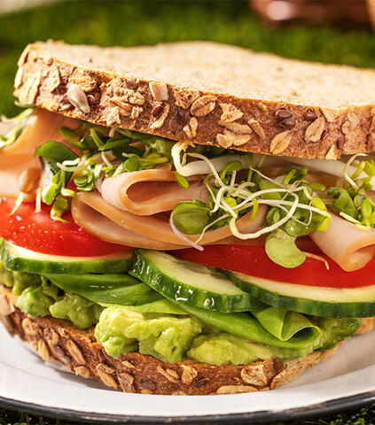 A close-up of a multigrain bread sandwich, filled with deli meats, avocado, and various vegetables &amp; sprouted grains