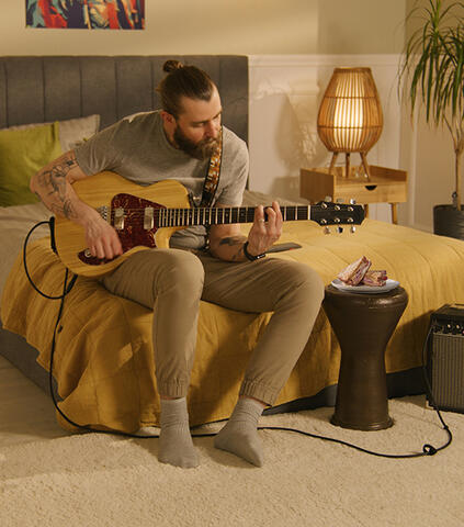 Man with long hair wrapped in a bun, sitting on the edge of a bed, playing the guitar